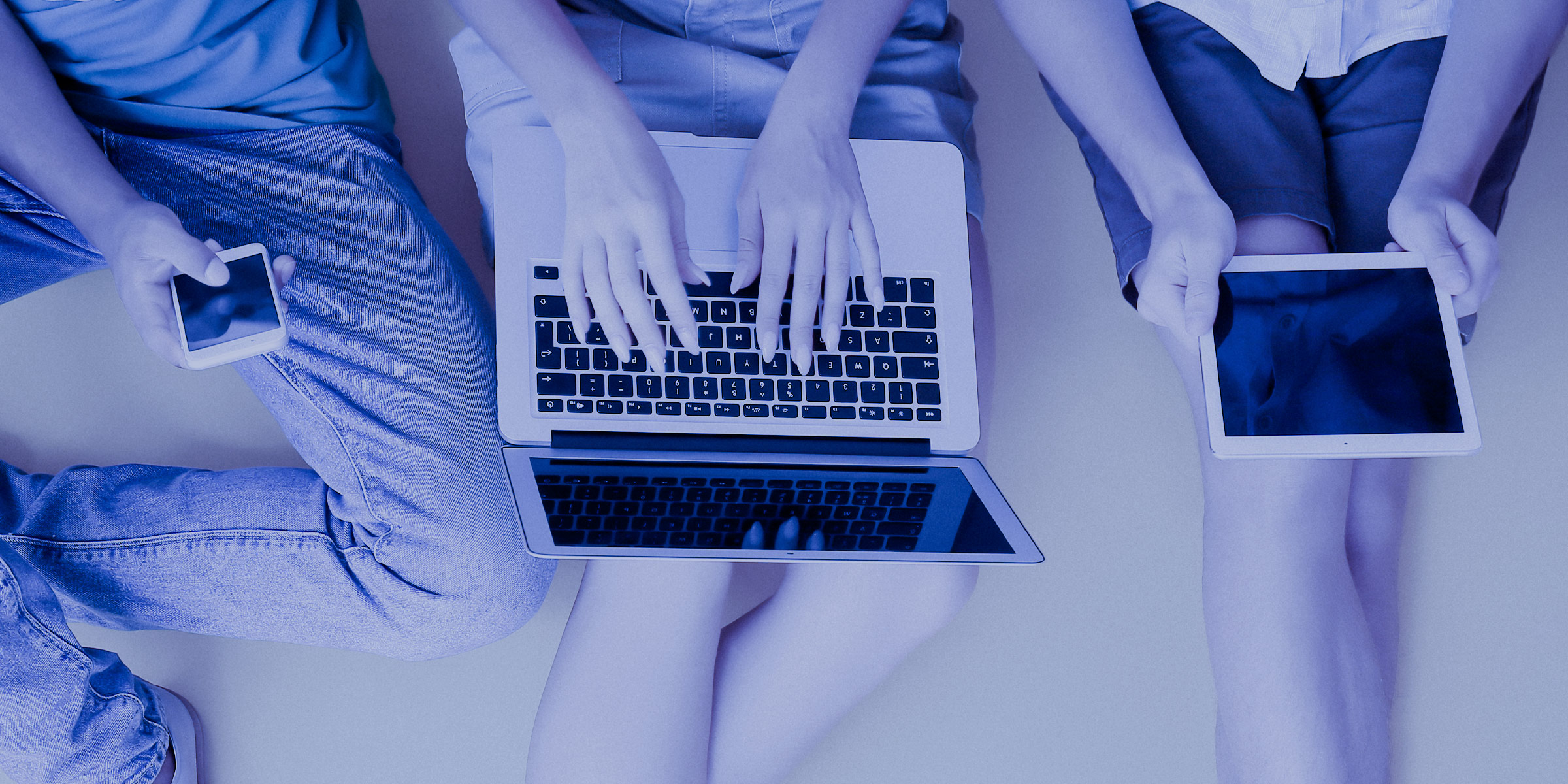Image from above of three people, one using a mobile phone, one using a laptop computer, and the other using a tablet
