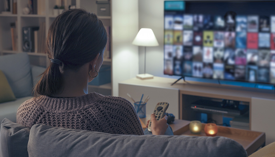 Over-the-shoulder view of woman holding smart TV remote while watching tv in dimly lit room